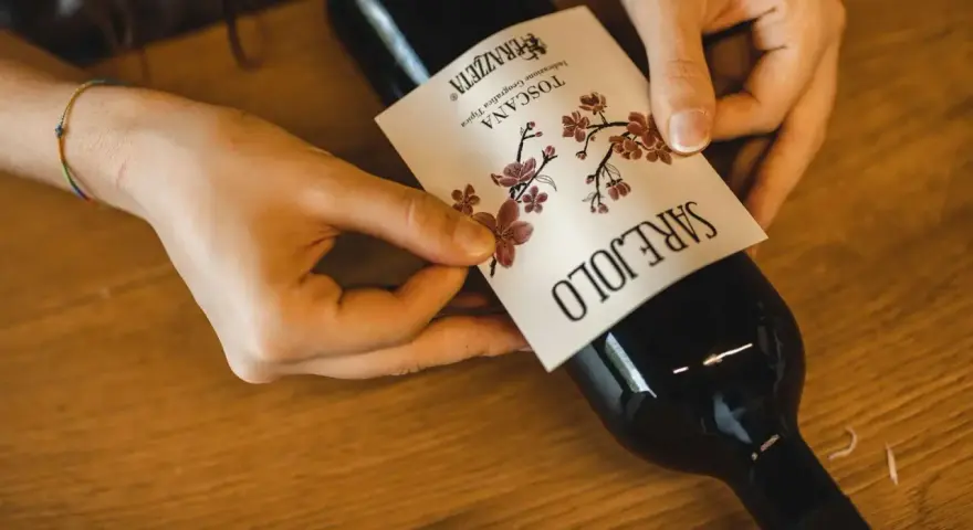 Mani che applicano un'etichetta su una bottiglia di vino. L'etichetta indica "Toscana", la regione di origine del vino Sangiovese.
