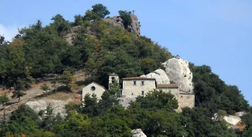 Antico eremo di Cerbaiolo, un complesso di edifici in pietra arroccato su un colle boscoso, con una croce sulla cima di una roccia.