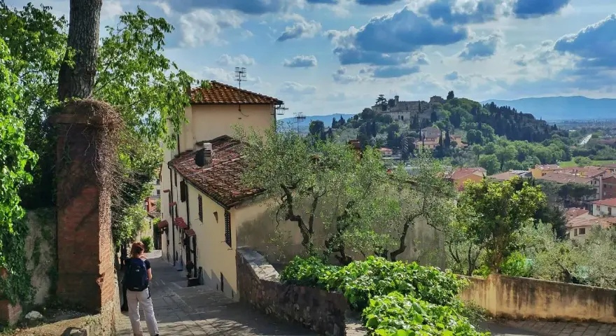 Una persona scende una scalinata in pietra, con case antiche e vegetazione rigogliosa. In lontananza, il **monte Morello** domina il paesaggio collinare.