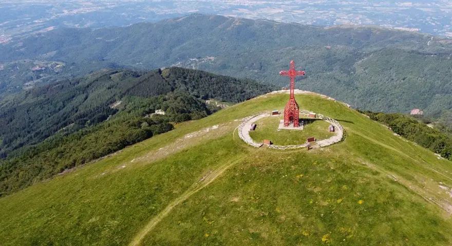 La croce del Pratomagno domina un paesaggio verdeggiante, con montagne boscose e una città in lontananza.
