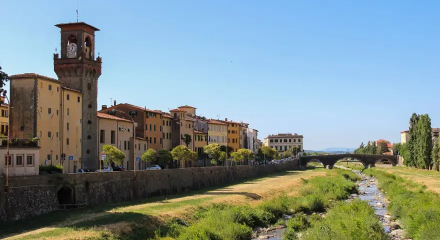 Pescia: cosa vedere? Un fiume serpeggia tra case colorate e un'antica torre con orologio, sotto un cielo azzurro.