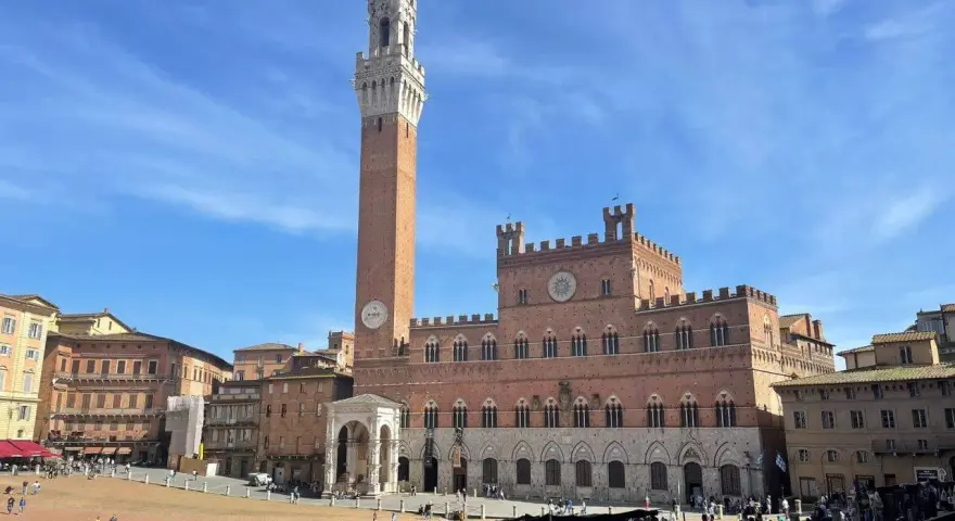 La Piazza del Campo di Siena, con la Torre del Mangia che domina il cielo azzurro, brulica di gente che passeggia e si gode il sole.