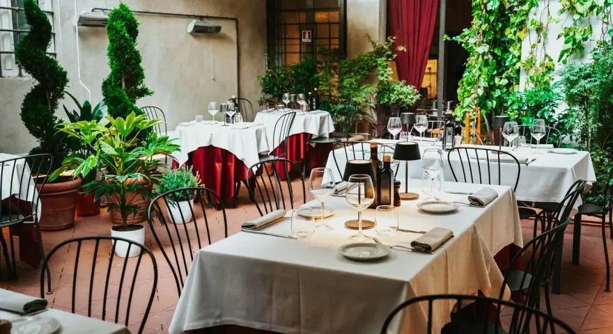 Cortile di un ristorante, tavoli apparecchiati con tovaglie bianche e rosse, piante verdi e sedie nere. Un invito a mangia e bevi santo spirito.