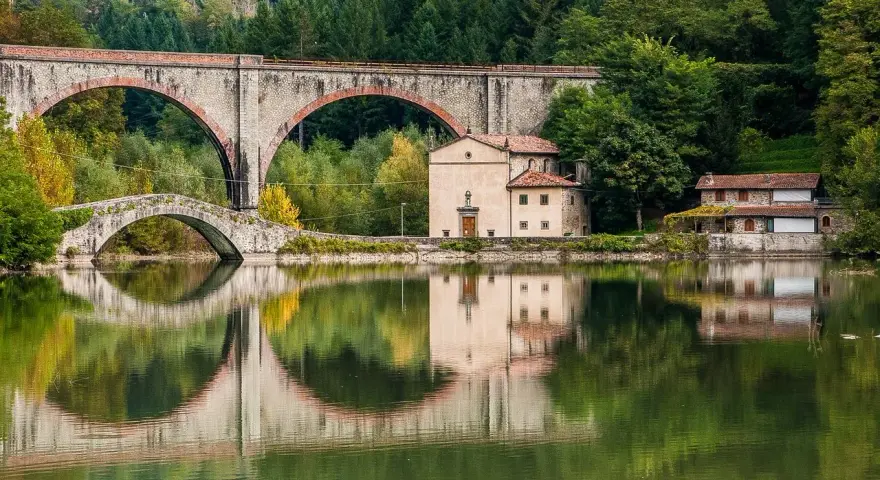 Pieve Fosciana: due ponti, uno antico e uno moderno, si specchiano in un lago calmo, con edifici storici e una foresta sullo sfondo.