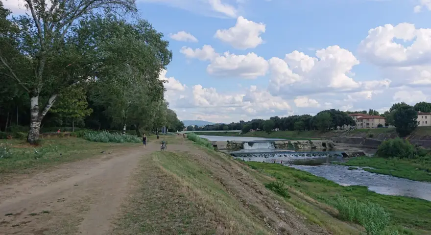 Sentiero lungo il fiume con alberi e il masso della Gonfolina in lontananza. Cielo azzurro con nuvole bianche.