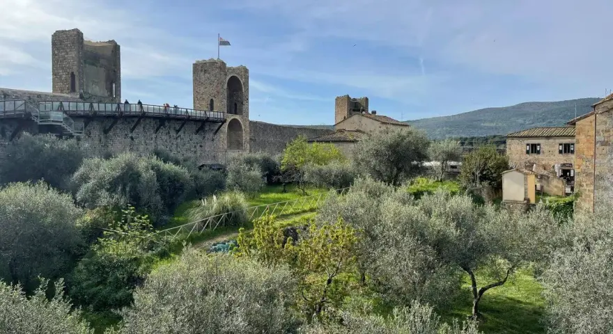 Le mura di Monteriggioni, Siena, dominano un paesaggio toscano con ulivi e colline. Un ponte pedonale attraversa la fortificazione.