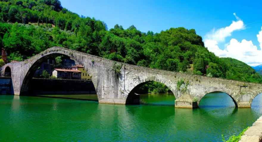Il Ponte del Diavolo, con i suoi archi in pietra, attraversa un fiume verde smeraldo, circondato da colline lussureggianti e un cielo azzurro.