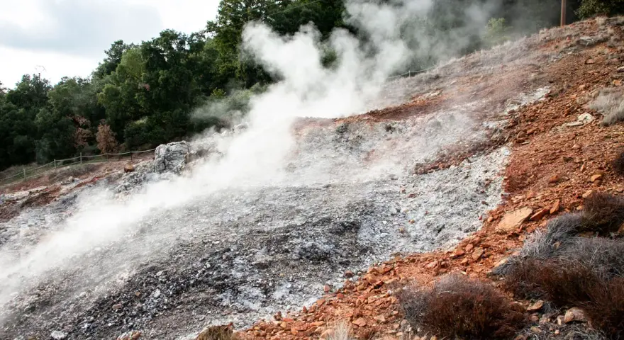 Fumarole attive nel Parco delle Biancane, con vapore che sale da un terreno roccioso e argilloso, circondato da vegetazione e alberi.