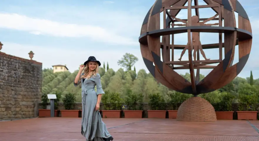 Donna con cappello nero ammira l'Uomo Vitruviano in una sfera di legno, un'opera iconica da vedere a Vinci, con colline verdi sullo sfondo.
