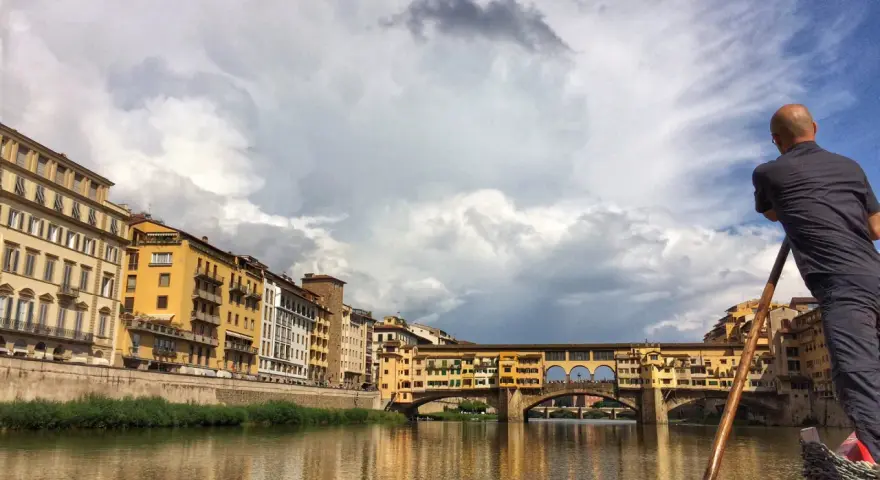 Un uomo su una barca a remi naviga lungo l'Arno, ammirando Ponte Vecchio e gli edifici storici di Firenze.