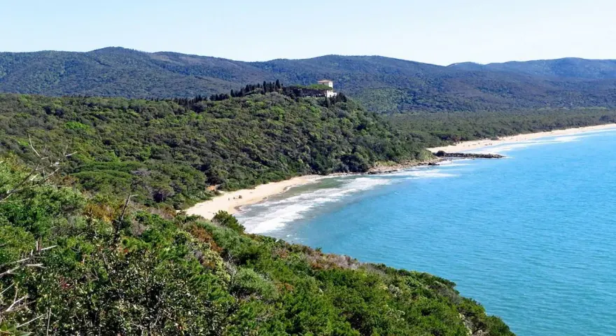 Vista panoramica delle bandite di Scarlino, con una spiaggia dorata bagnata da un mare azzurro e una villa in cima a una collina verde.