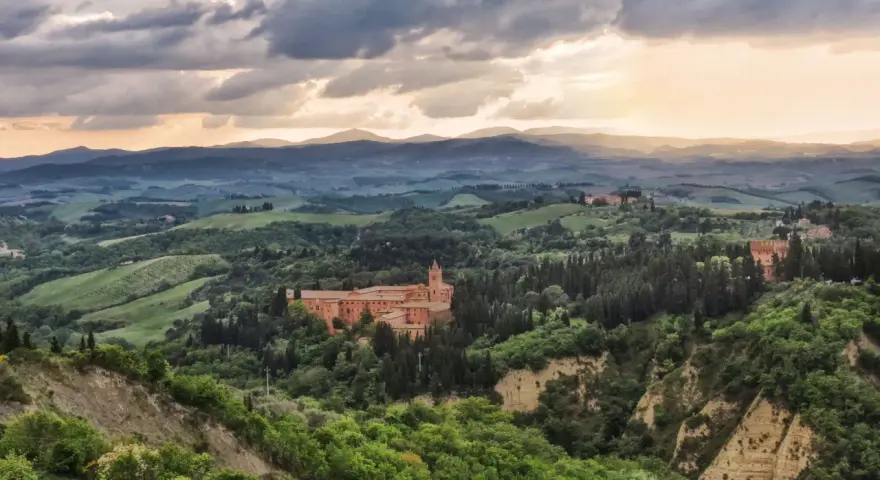 Un imponente complesso monastico, il Monte Oliveto Maggiore, si erge tra dolci colline toscane sotto un cielo drammatico.