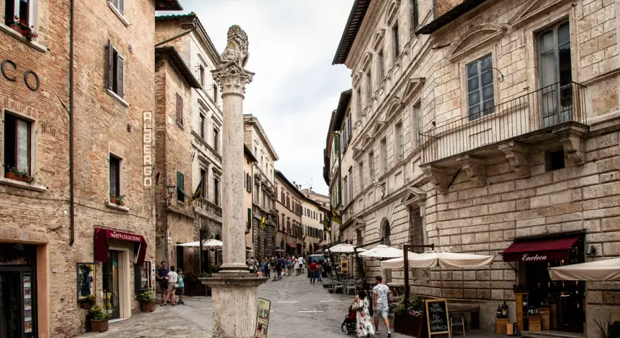 A stone column with a lion statue stands in a cobblestone street lined with historic buildings and shops.