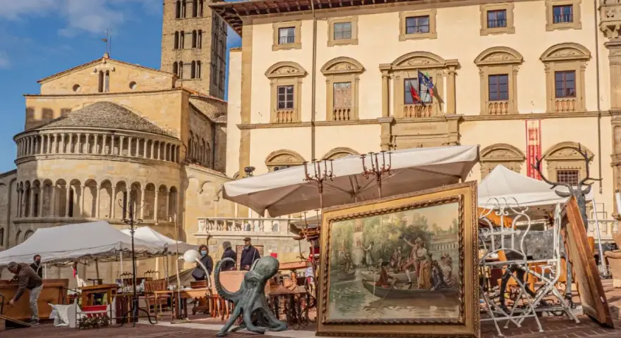 Mercatino dell'antiquariato in piazza grande, con opere d'arte, sculture e mobili antichi esposti sotto il sole.