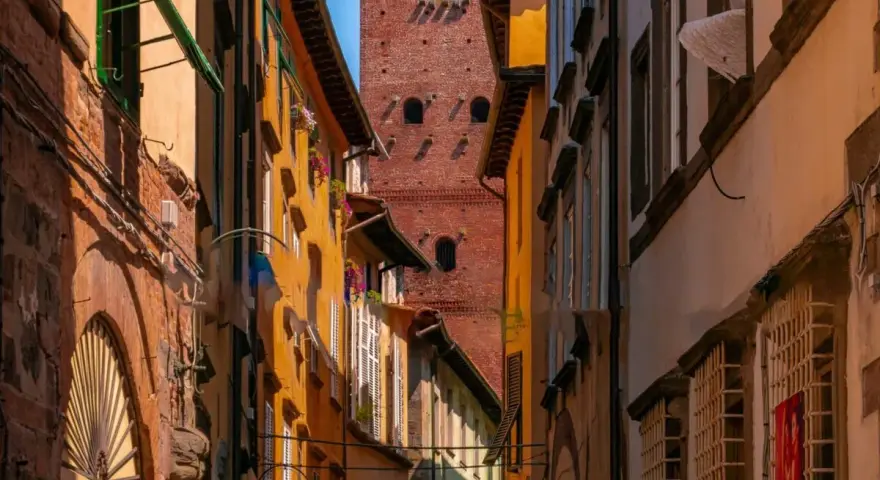 Un vicolo stretto a Lucca con edifici colorati e la torre Guinigi che svetta in lontananza.