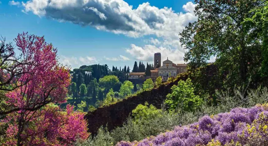 Giardini fioriti con glicini viola e alberi rosa, sullo sfondo un edificio storico con torre: curiosità da vedere a Firenze.