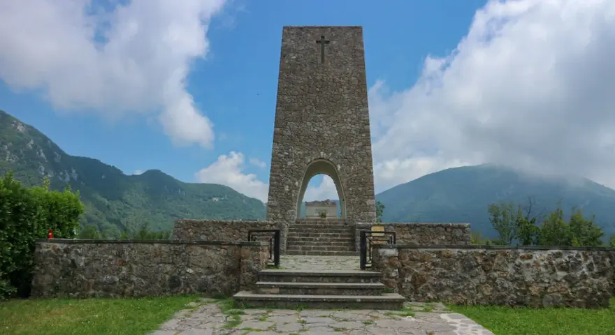 Monumento a Sant'Anna di Stazzema, cosa vedere: un imponente torrione in pietra con arco, che custodisce una tomba, circondato da mura e montagne.