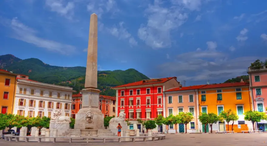 Piazza con obelisco e leoni, edifici colorati e montagne sullo sfondo. Un'idea di cosa vedere a Marina di Massa.