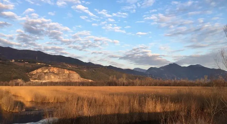 Acque calme del lago di Porta riflettono un cielo azzurro con nuvole bianche, circondato da canneti dorati e montagne all'orizzonte.