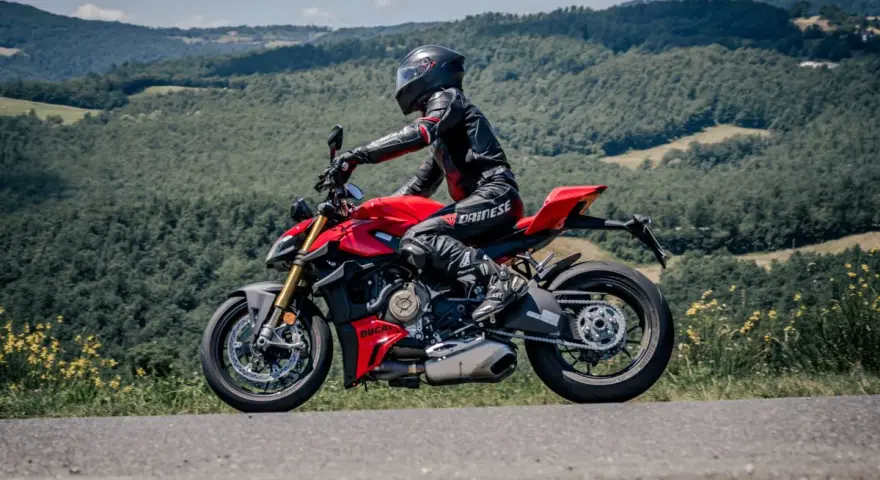 Motociclista in tuta rossa e nera sfreccia su una Ducati rossa lungo il passo del giogo, tra colline verdi e cielo azzurro.