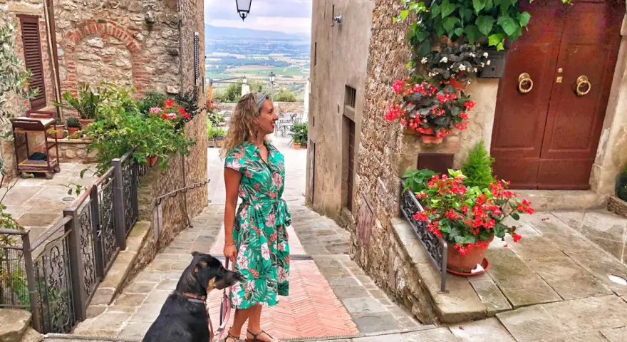 Una donna con un cane passeggia in un vicolo fiorito di Civitella Val di Chiana, con una vista panoramica sullo sfondo.