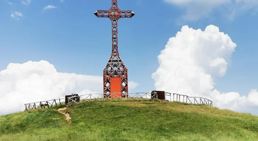 Croce metallica rossa sulla cima di una collina erbosa, con un sentiero che sale. Il **Pratomagno Arezzo** domina il paesaggio sotto un cielo azzurro con nuvole bianche.