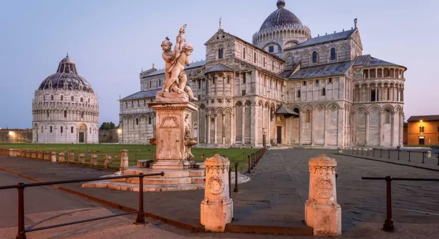 Il **Battistero di Pisa** e il Duomo illuminati dalla luce dell'alba. Una fontana con putti al centro della piazza.