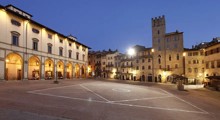 Piazza illuminata di Arezzo al crepuscolo, con portici, negozi e una torre medievale. Qui la vita è bella.