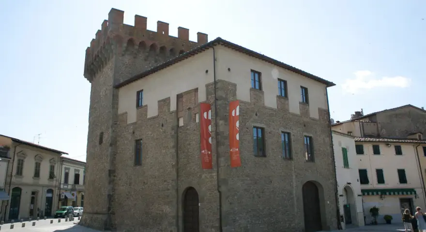 Il cassero di Montevarchi, un'antica torre con merlature, si erge imponente accanto a edifici storici, con bandiere rosse che adornano la facciata.