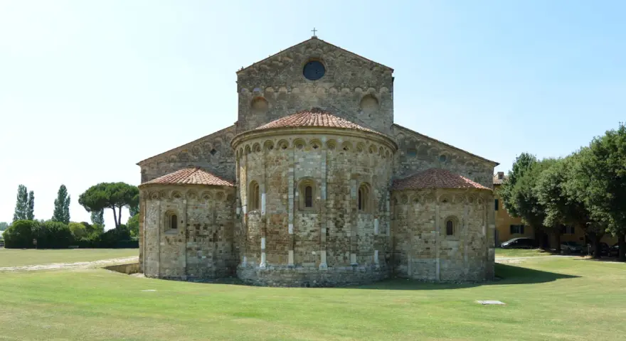 La Basilica di San Piero a Grado, con le sue absidi romaniche e il prato verde circostante, si staglia contro un cielo limpido.