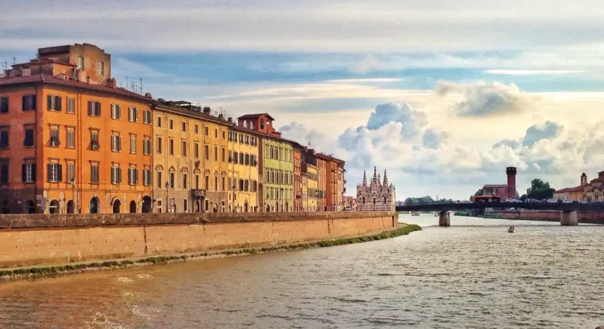 Edifici colorati lungo il fiume Arno a Pisa, con un ponte e nuvole suggestive nel cielo. Un'immagine che cattura la bellezza di Pisa e dintorni.