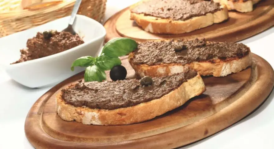 Toasted bread slices topped with dark spread, garnished with basil, olives, and capers.