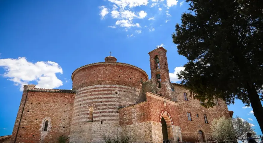 L'abbazia di San Galgano, con la sua rotonda in mattoni a fasce bianche e rosse, si staglia sotto un cielo azzurro con nuvole bianche.