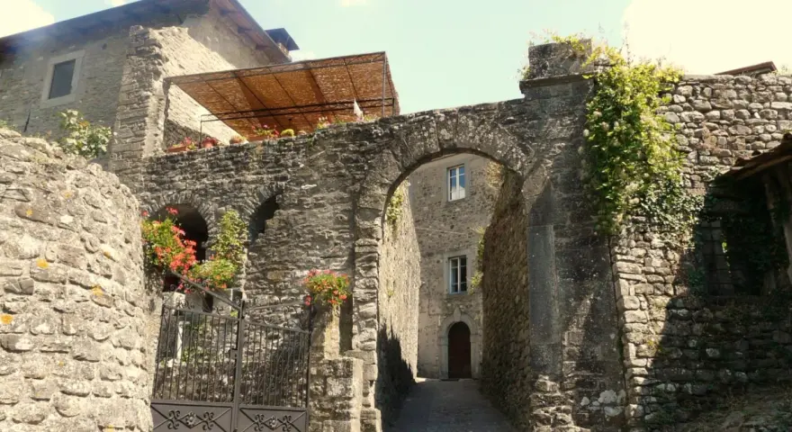 Un vicolo antico a Casola in Lunigiana, con un arco in pietra, mura di sassi e fiori rossi che sbocciano.