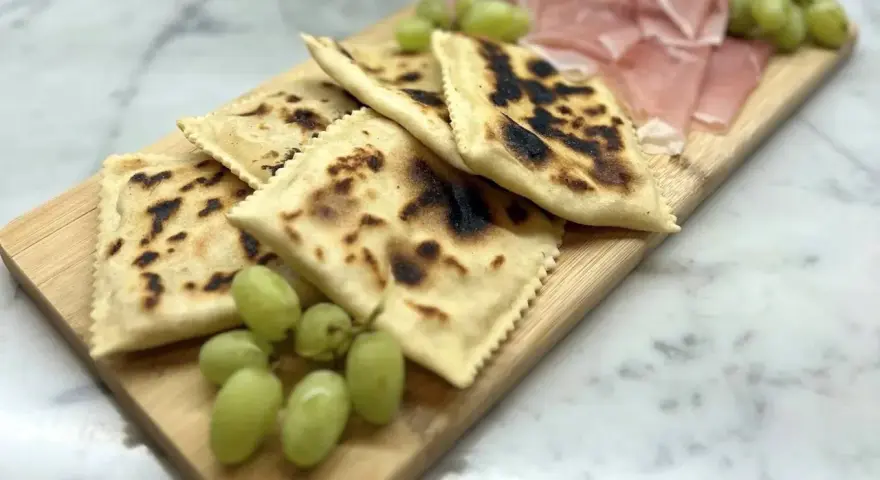 Tortelli alla lastra casentino, dorati e leggermente bruciacchiati, serviti con uva fresca e affettati di prosciutto su un tagliere di legno.
