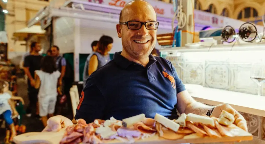 Uomo sorridente al mercato delle vettovaglie Livorno, offre un tagliere di salumi e formaggi.