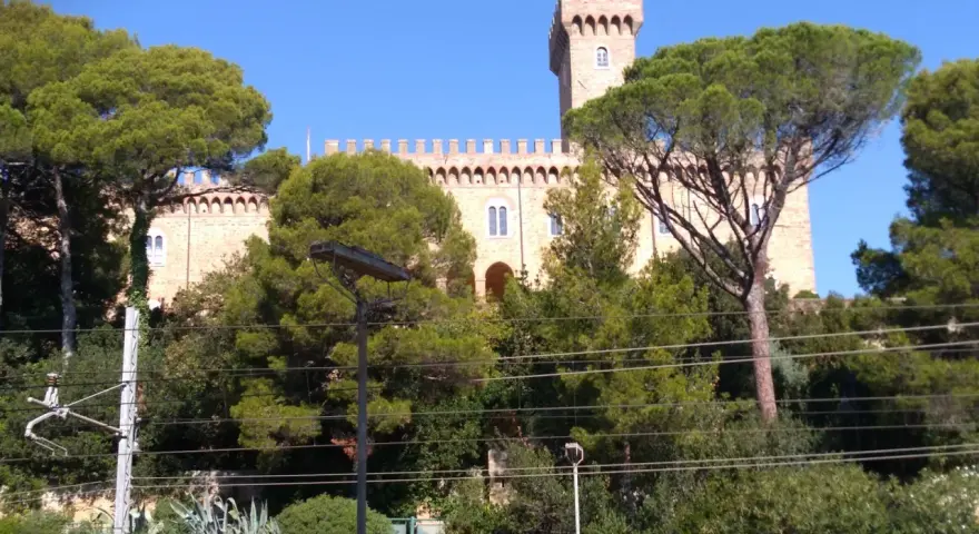 Il Castello Pasquini, con la sua torre imponente, emerge tra alberi verdi e un cielo azzurro.