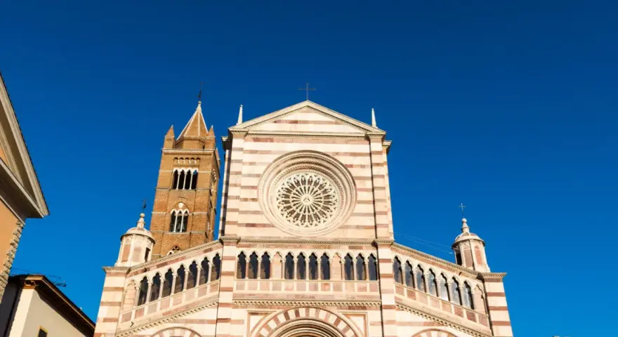 Il Duomo di Grosseto, con la sua facciata a strisce bianche e rosa e il campanile in mattoni, si staglia contro un cielo azzurro intenso.