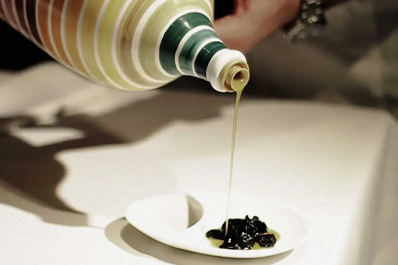 Un filo d'olio d'oliva dorato scende da una bottiglia decorata su un piatto con olive nere, per un'esperienza di degustazione.