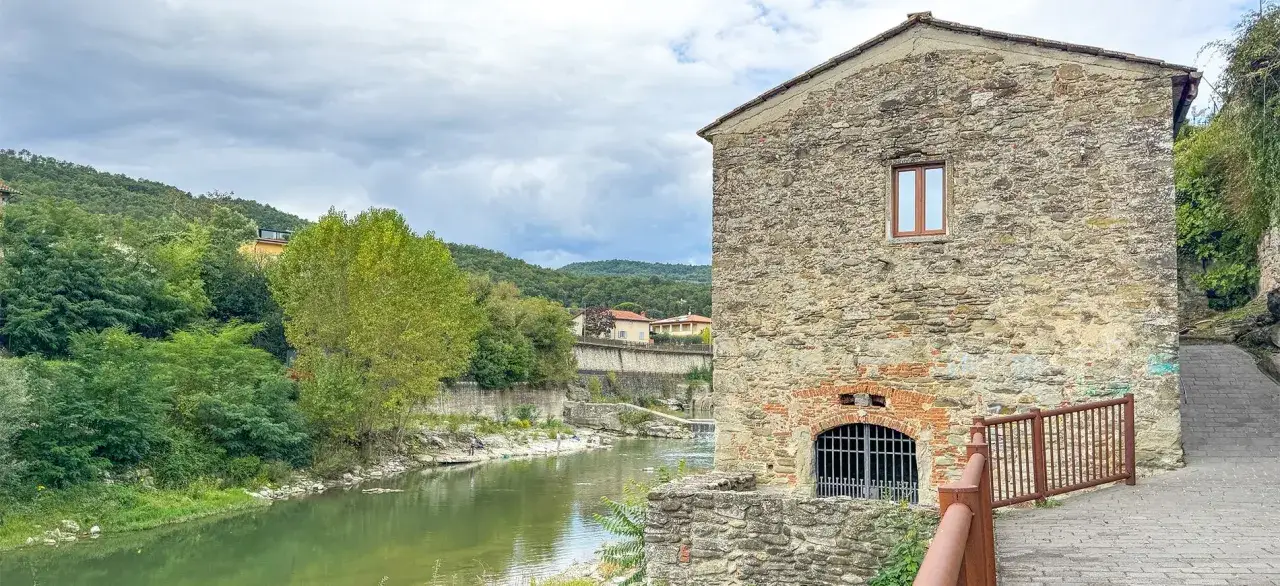Antico edificio in pietra a Subbiano, Arezzo, con un fiume e colline verdi sullo sfondo.