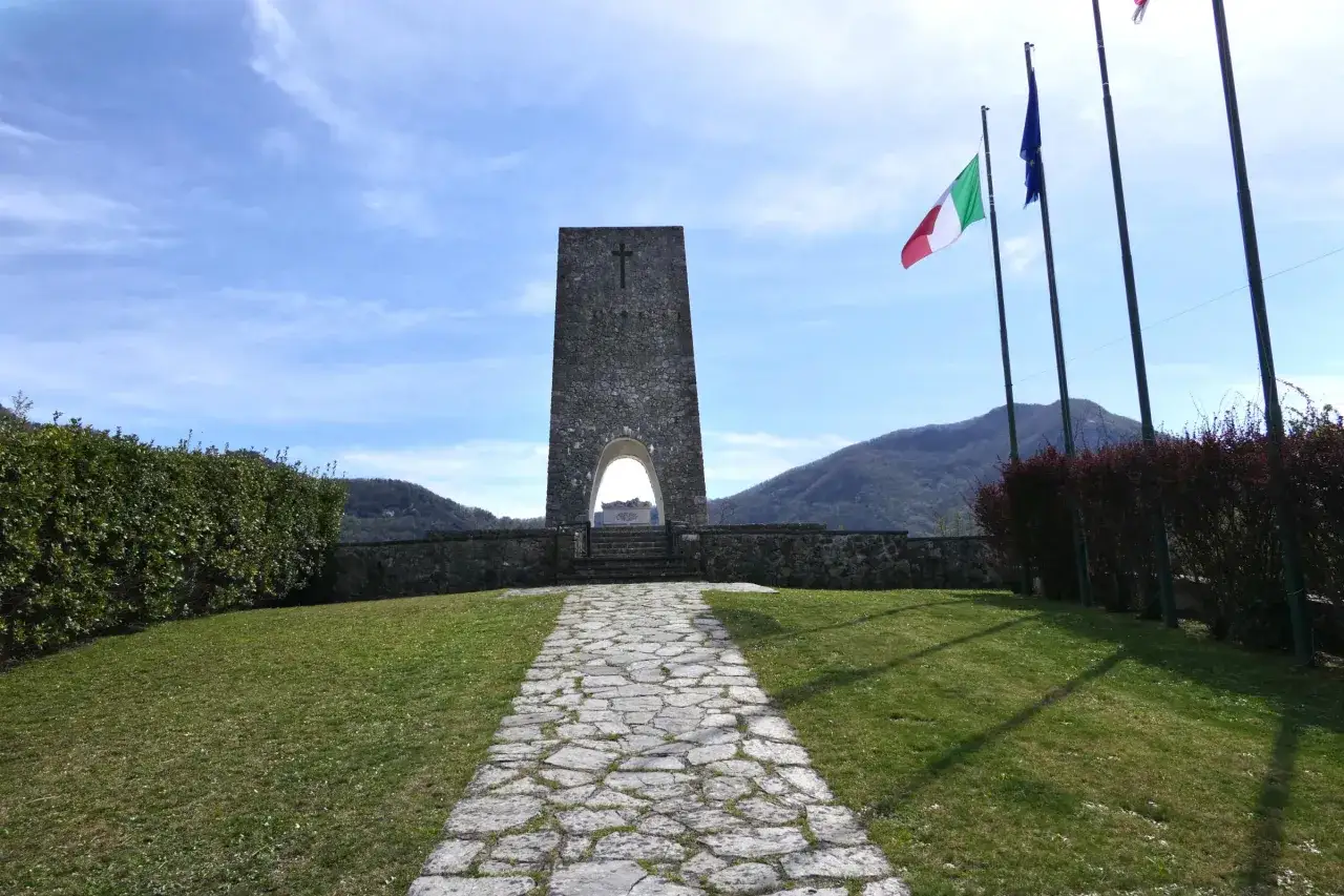 Monumento a Sant'Anna di Stazzema, cosa vedere: un'imponente torre in pietra con una croce, un arco e un sentiero lastricato che conduce verso le montagne.