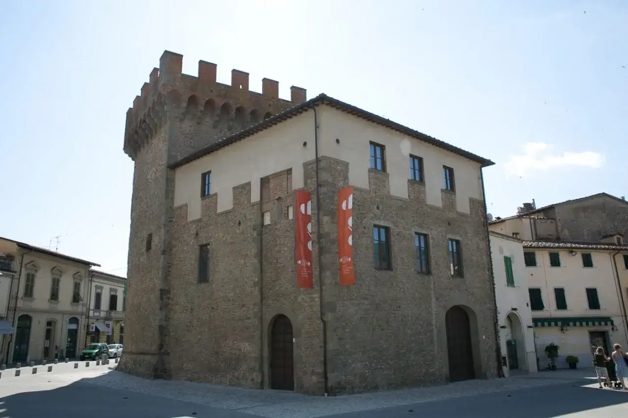 Il cassero di Montevarchi, un'antica torre con merlature, si affaccia su una piazza con edifici storici e bandiere arancioni.