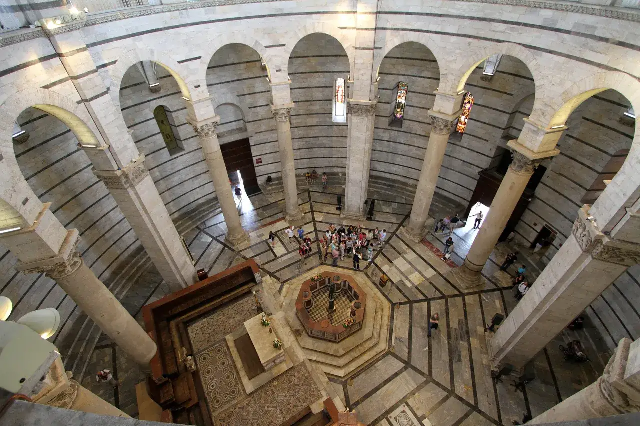 Interno del Battistero di Pisa, con colonne, archi e un gruppo di turisti attorno alla vasca ottagonale.