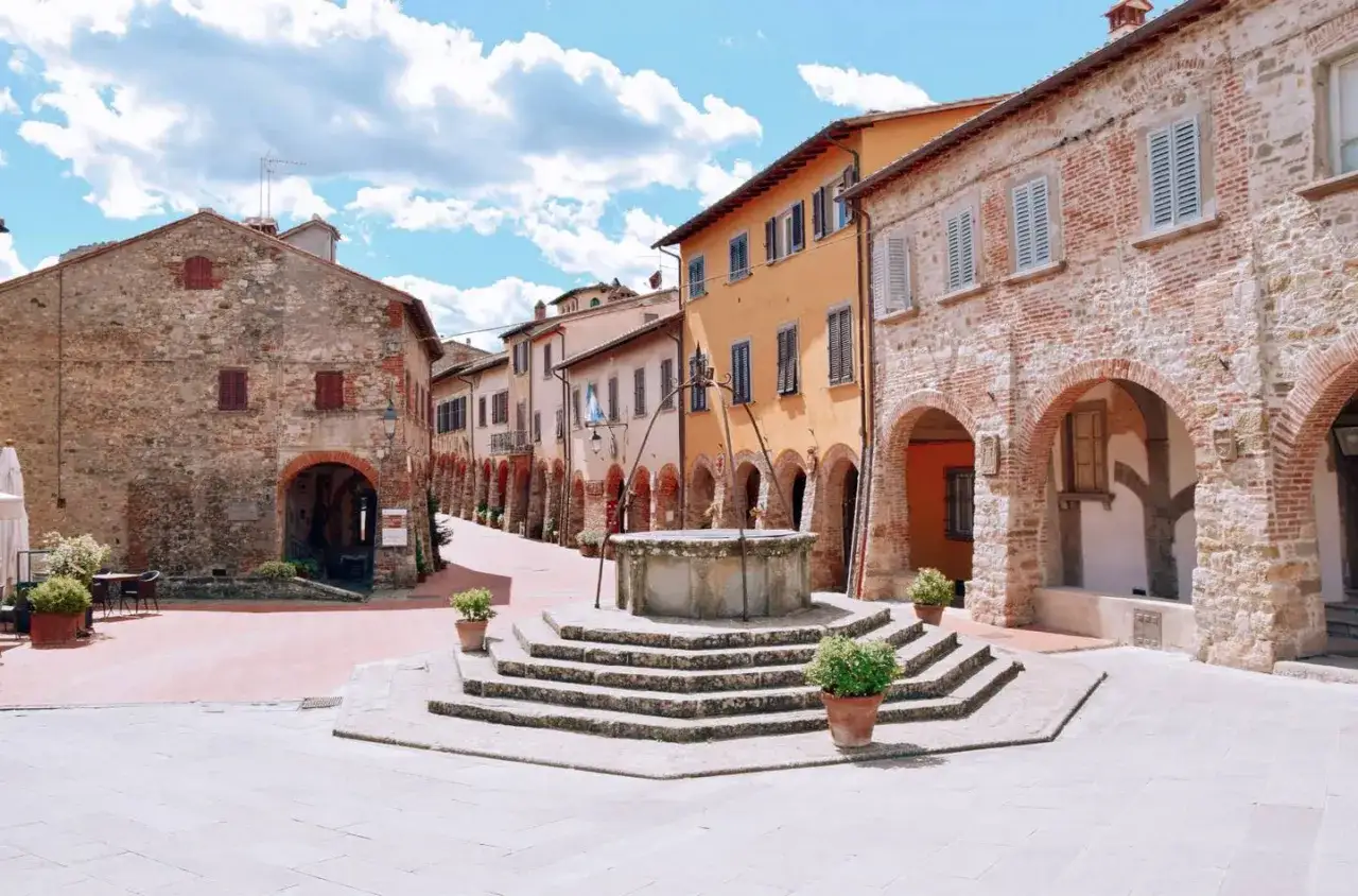 Piazza con pozzo antico a Civitella Val di Chiana, circondata da case in pietra e mattoni con portici e finestre con persiane.