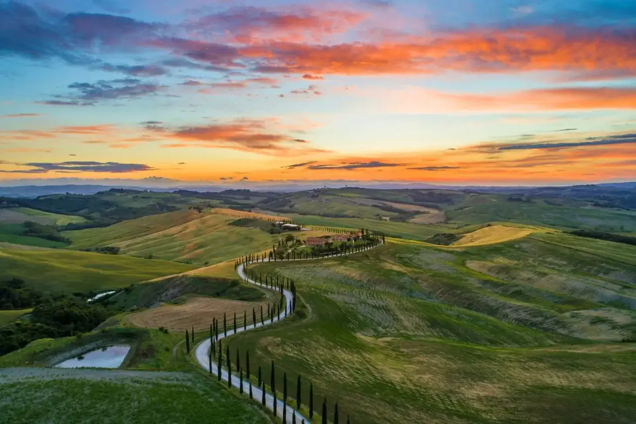 Dolce tramonto sulle colline toscane, con la Via Francigena che serpeggia tra cipressi e campi verdi.