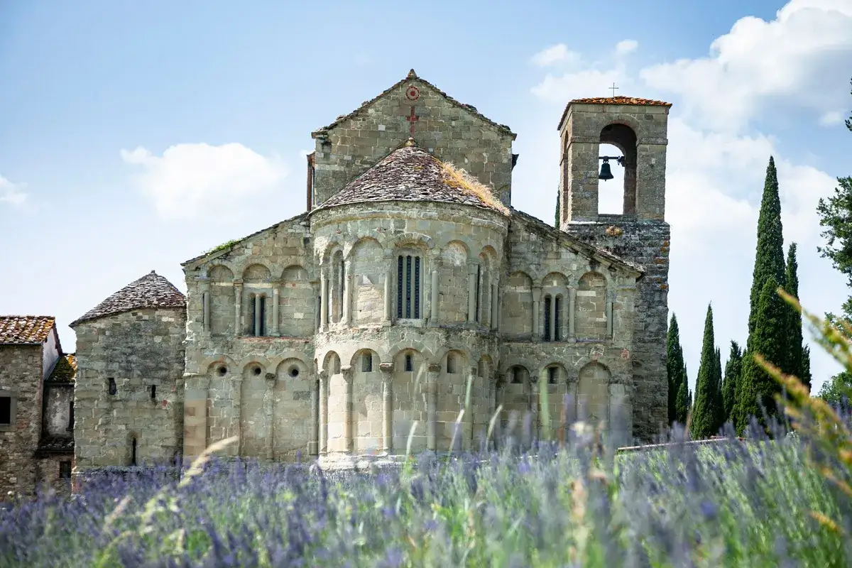 Antica chiesa romanica, con un campanile e cipressi, immersa in un campo di lavanda. Un'immagine di serena accoglienza.