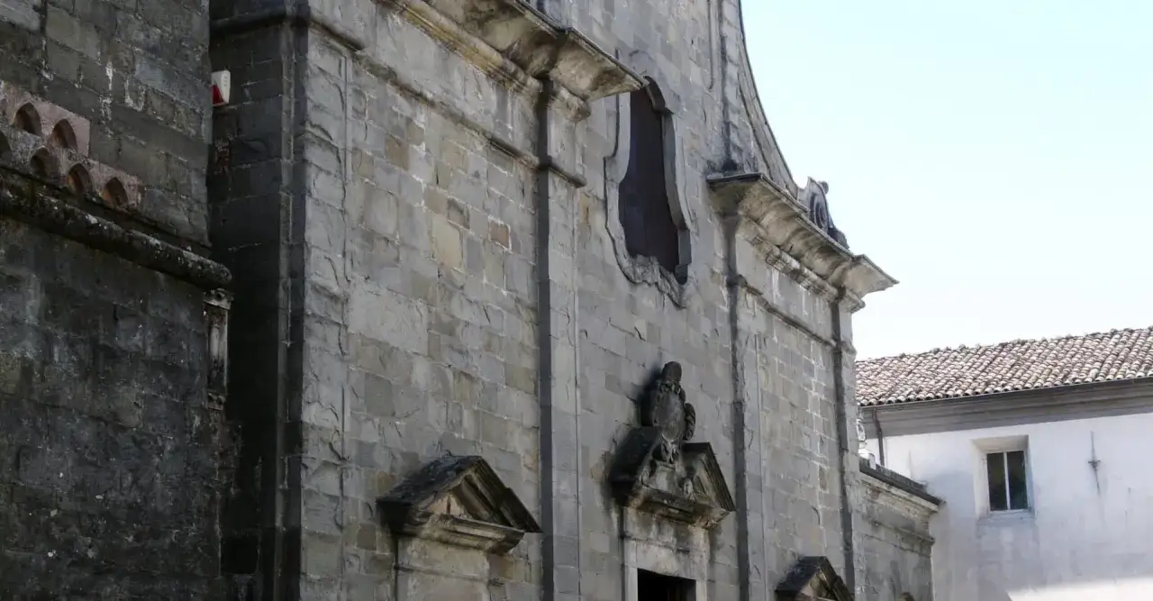 Facciata in pietra di una chiesa a Pieve Fosciana, con un'antica finestra e un bassorilievo.