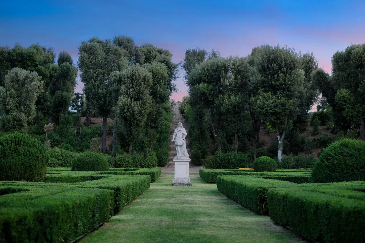 Giardino all'italiana a San Quirico d'Orcia con labirinto di siepi, statua e viale alberato al tramonto.