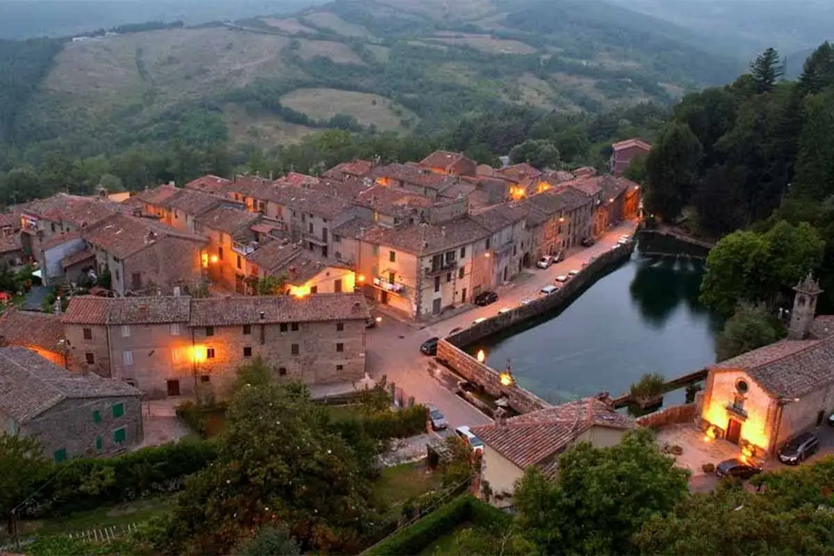 Incantevole vista serale di Santa Fiora, con le case illuminate che si affacciano su un laghetto tranquillo, circondato da colline verdi.