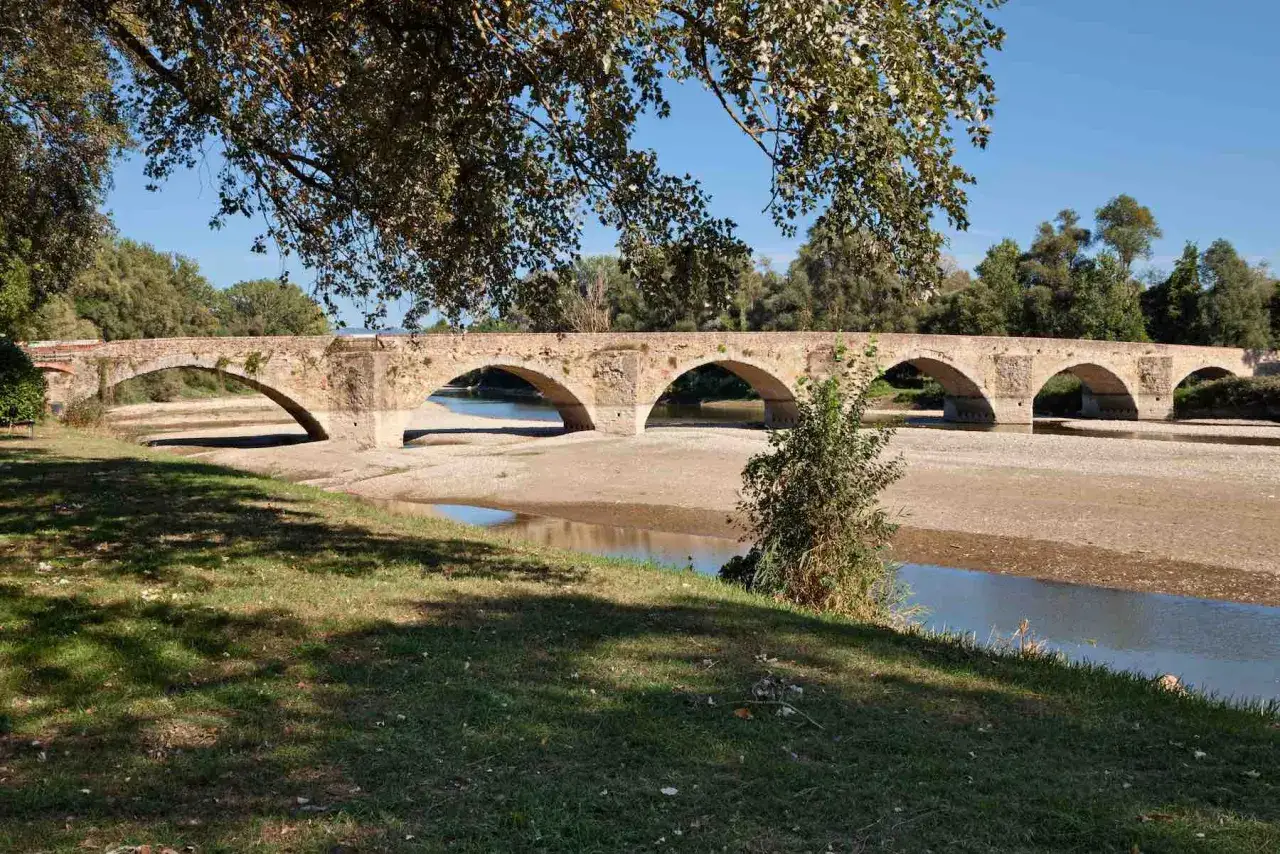 Il Ponte Buriano, con i suoi archi in pietra, attraversa un fiume con poca acqua, circondato da alberi e vegetazione lussureggiante sotto un cielo azzurro.